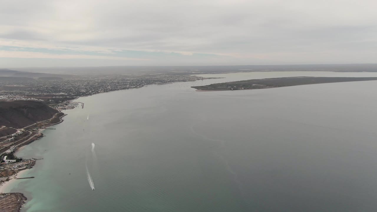 Drone view of the La Paz waterway with a pale boat trail near the center-left water, a curved shoreline and hills to the right, distant urban area, and flat land across the horizon, with no visible people.