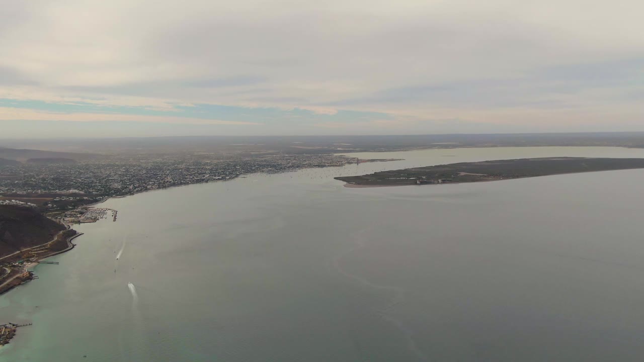 Wide aerial frame over La Paz, Baja California Sur, showing green-gray bay water, city shoreline, a low landmass across the water, cloudy sky, and no visible people.