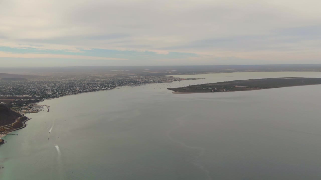 Aerial view over La Paz, Baja California Sur, with the city along the shore, open bay water in front, a white boat wake lower in frame, distant land, and no visible people.