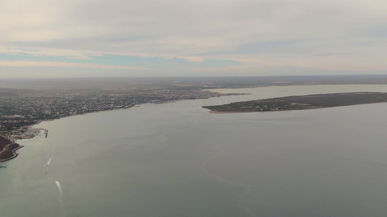 High aerial frame of La Paz, Baja California Sur, with calm bay water, the city edge, a small projecting shoreline area, a boat wake near the lower center, and no visible people.