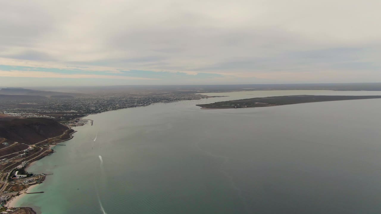 Aerial view over La Paz, Baja California Sur coastal waters, with pale shallows near shore, gray water farther out, white boat wakes, distant city, and no visible people.