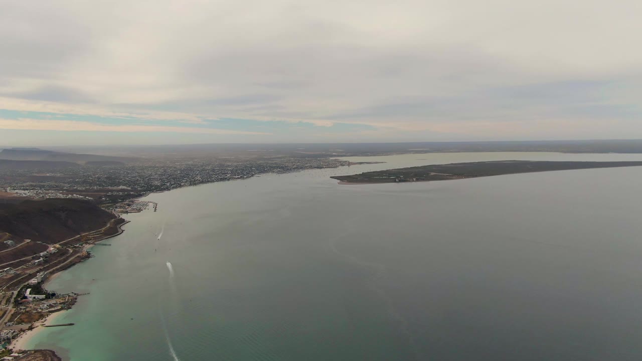 La Paz aerial frame showing boat wakes farther across the coastal water, with the curved shore and low dark hill at left, distant city on the horizon, and no visible people.