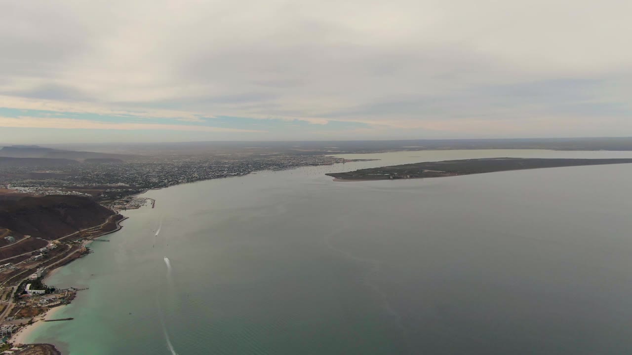 Wide drone view above La Paz coastal water, with a broad gray surface in the foreground, a thin white wake near shore, city blocks beyond the shoreline, and no visible people.