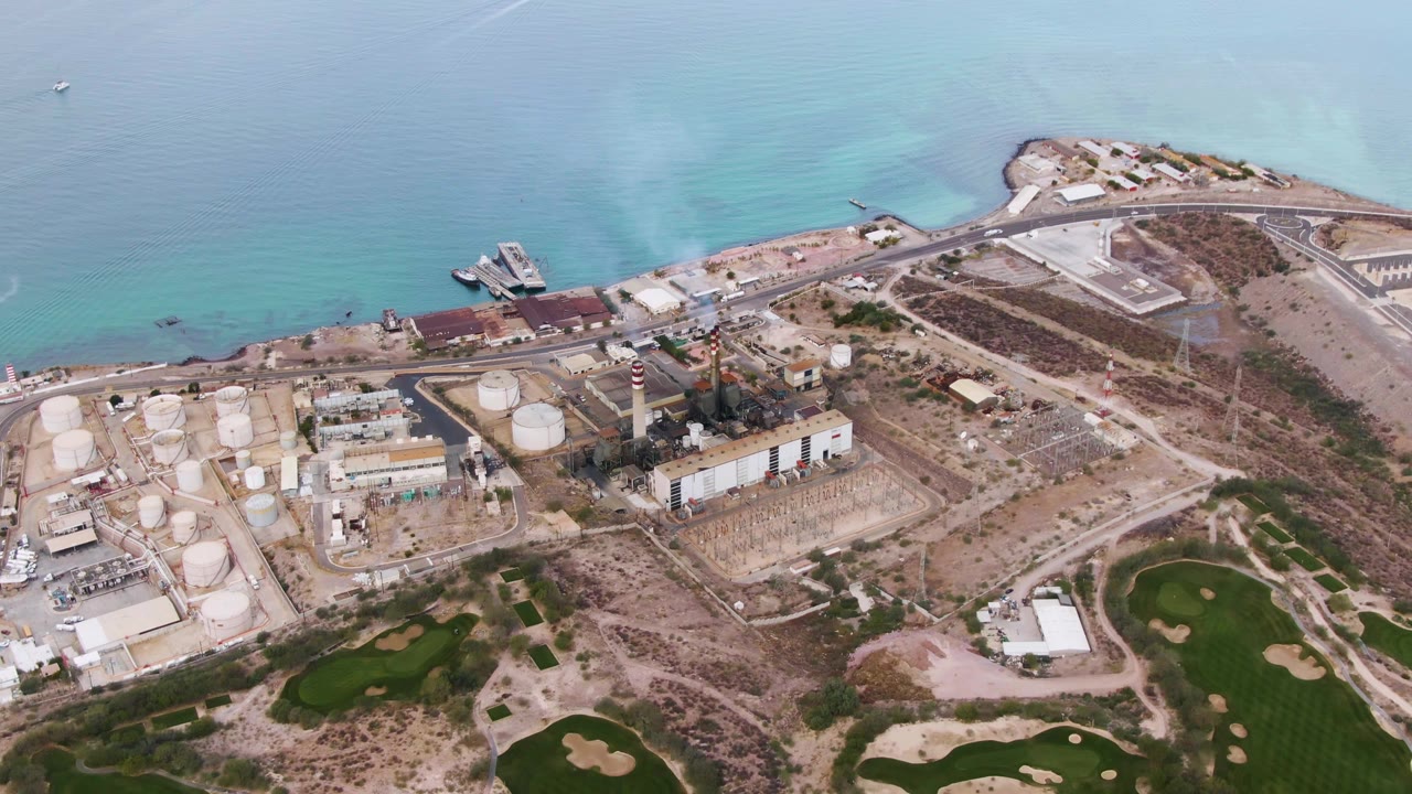 Opening aerial frame over La Paz, Baja California Sur, showing pale blue water, shoreline facilities, storage tanks, industrial buildings, sandy lots, and no visible people.