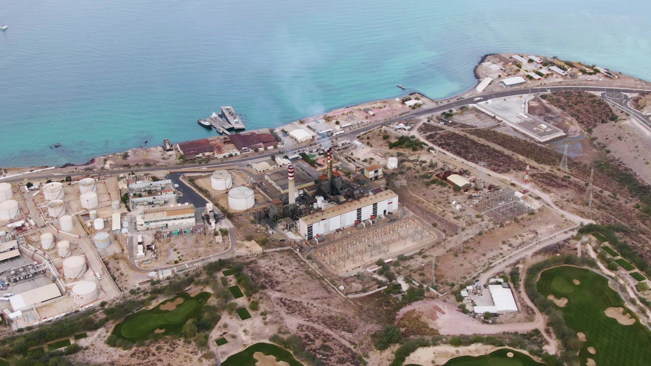 Second aerial frame in La Paz, Baja California Sur, with the tank farm and striped stack centered below the turquoise shallows, plus golf-course green patches at the lower edge and no visible people.