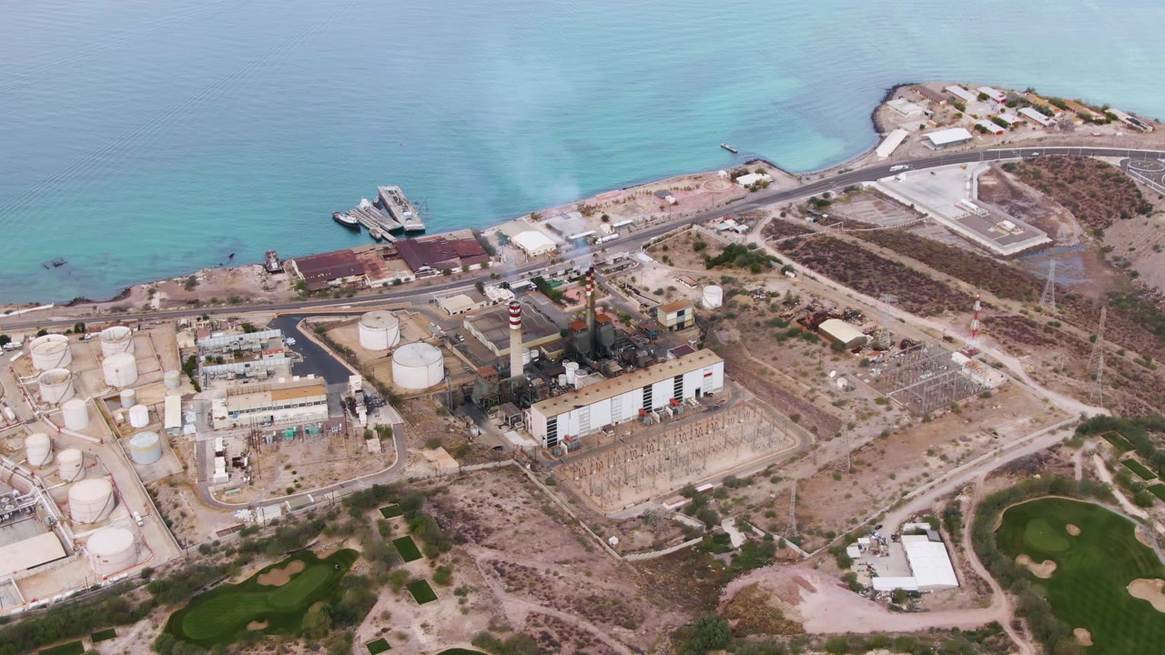 Third frame over La Paz, Baja California Sur, showing white storage tanks on the left, low industrial buildings in the center, arid open ground to the right, blue-green ocean beyond, and no visible people.
