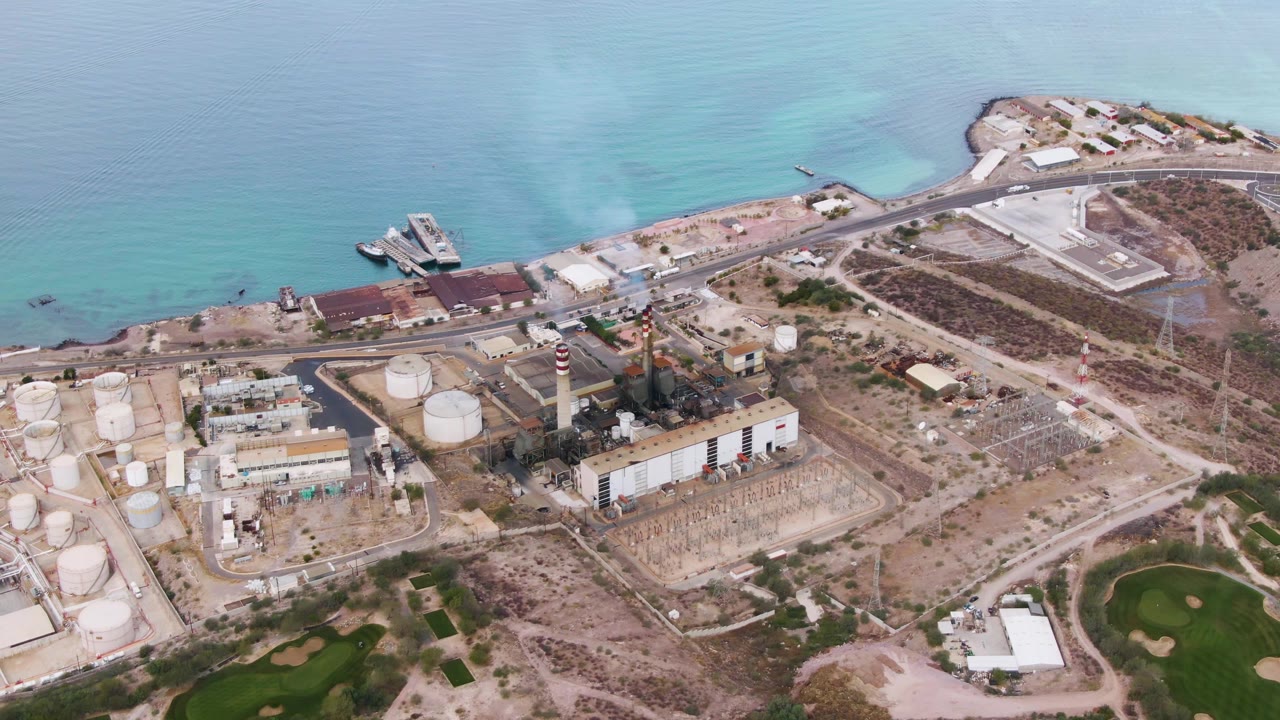 Coastal industrial complex beside ocean and golf-course green patches from above.