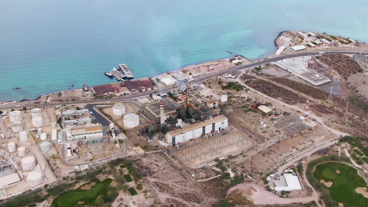Aerial frame in La Paz, Baja California Sur, showing the smoking stack and industrial buildings more centrally, with tank farms to the left, a curved shoreline road above, blue water beyond, and no visible people.