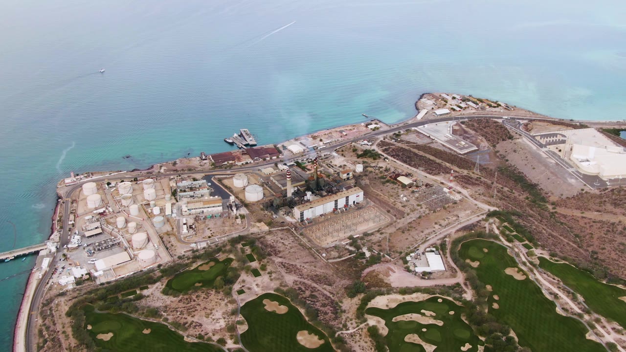 Aerial frame over La Paz, Baja California Sur, with white tanks and low industrial buildings near the shoreline, pale turquoise water behind them, green fields in the lower frame, and no visible people.