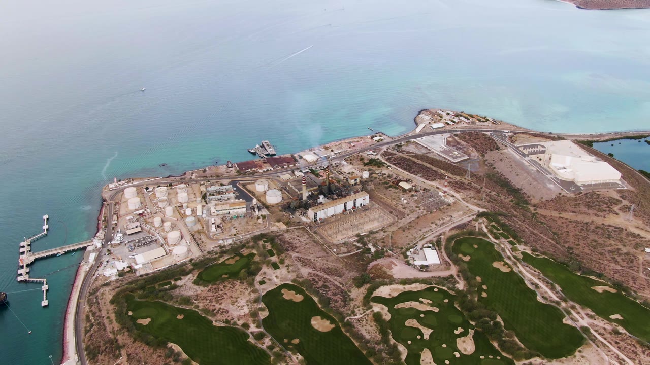 Mid-clip aerial frame over La Paz, Baja California Sur, showing the left-side dock, clustered tanks, low buildings, dry open land, green fields with pale patches, turquoise water, and no visible people.