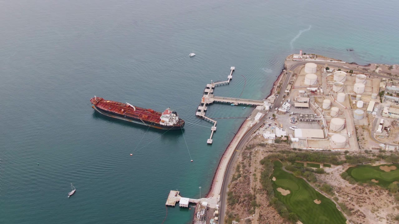 High aerial view over La Paz port facilities, with a red cargo ship on blue water, a white pier, shore tanks, a small vessel wake at the far left edge, and no visible people.