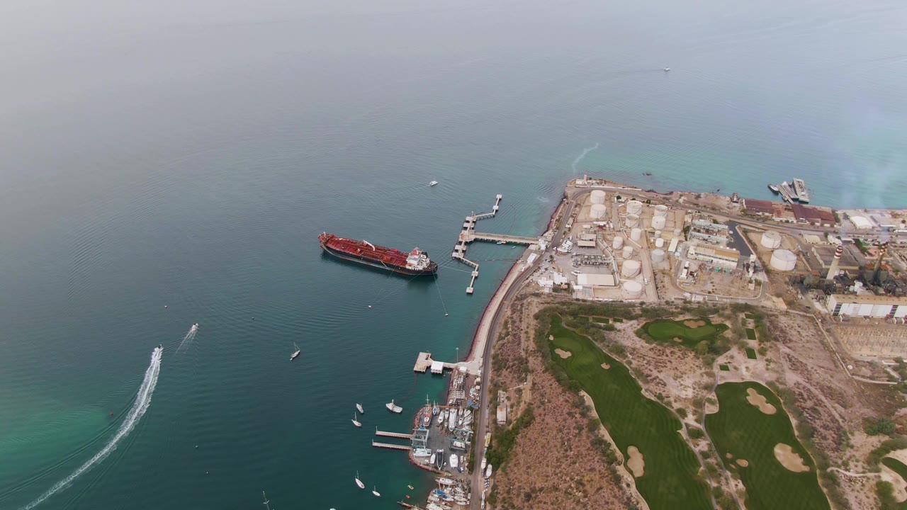 Aerial view over La Paz, Baja California Sur with teal water at left, a red ship near a pier, industrial tanks along the shore, green golf fairways, sand traps, and no visible people.