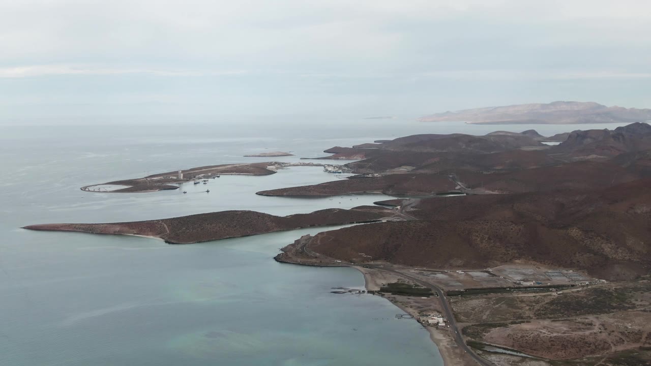 Aerial view in La Paz, Baja California Sur with the road and shoreline lower in frame, open bay water to the left, islands across the middle distance, and no visible people.