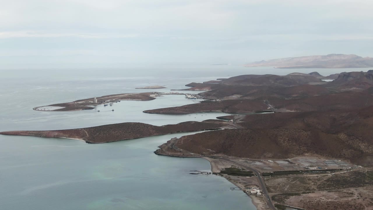 High aerial frame over La Paz, Baja California Sur showing the coastal roadway near the lower right, shallow turquoise water at left, island shapes offshore, and no visible people.