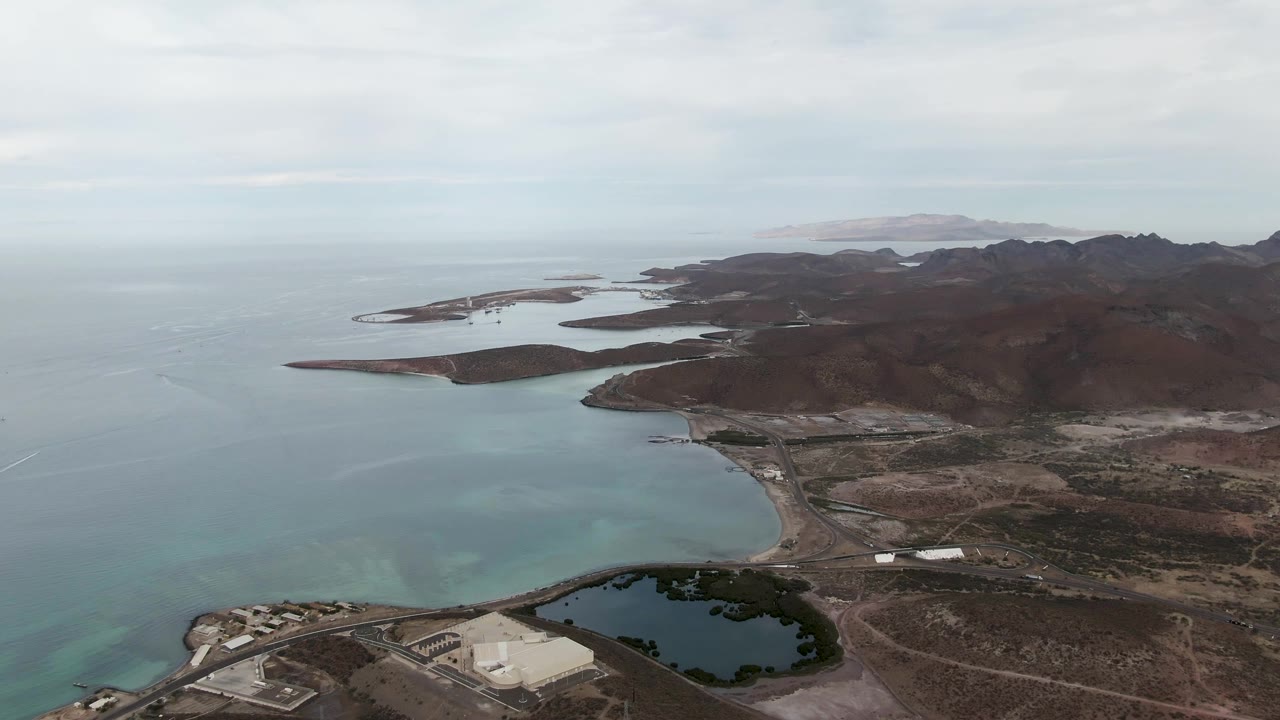 Aerial frame over La Paz showing more of the open bay at left, low islands in the distance, dry mountain shoreline at right, roads, a pond, and no visible people.