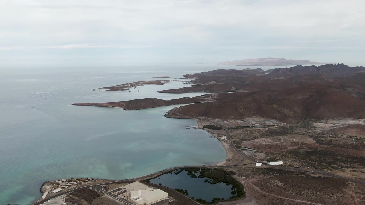 Aerial view of La Paz coastline with turquoise shallows in the bay, low islands along the horizon, dark mountain ridges, shoreline roads, a small pond, and no visible people.