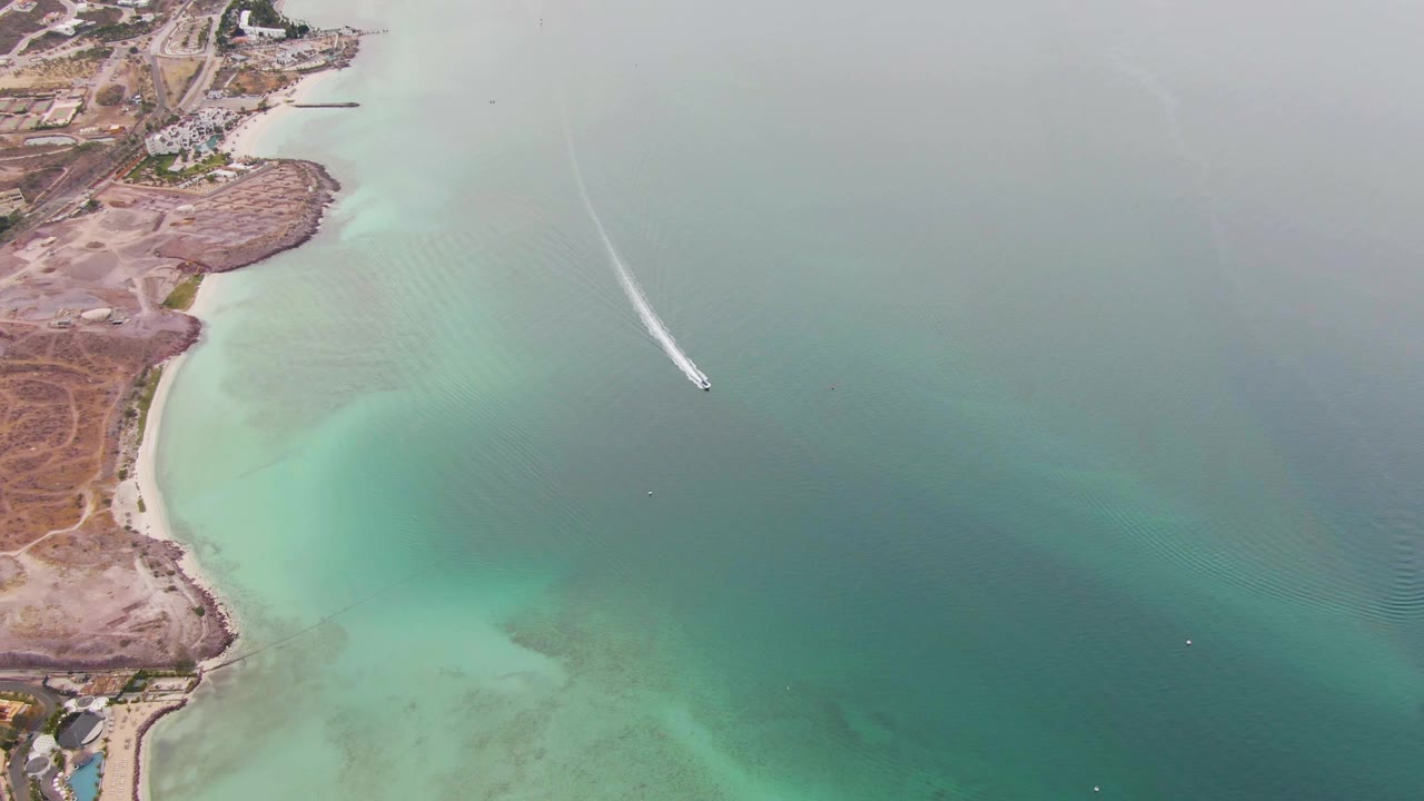 Aerial frame from La Paz, Baja California Sur showing a small boat crossing green-blue water near sandy and rocky shore, with no visible people.
