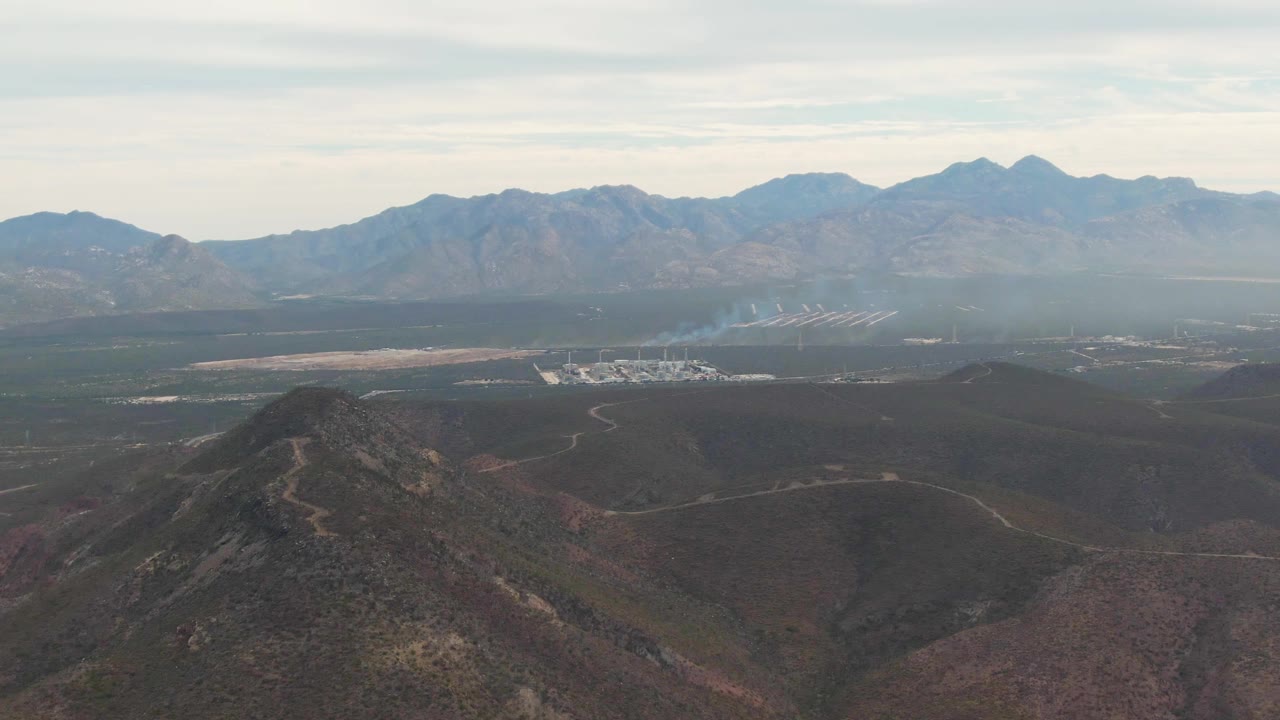 At 0:00, a La Paz, Baja California Sur aerial view shows a rocky foreground hill, broad valley floor, distant industrial buildings, a pale smoke plume, layered mountains, and no visible people.