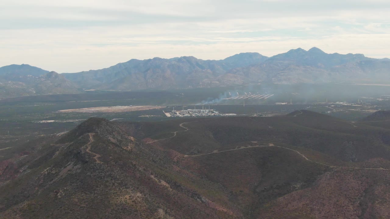At 0:02, the La Paz valley remains wide and still, with the foreground hill lower in frame, distant roads and facility structures visible beneath the smoke, and no visible people.