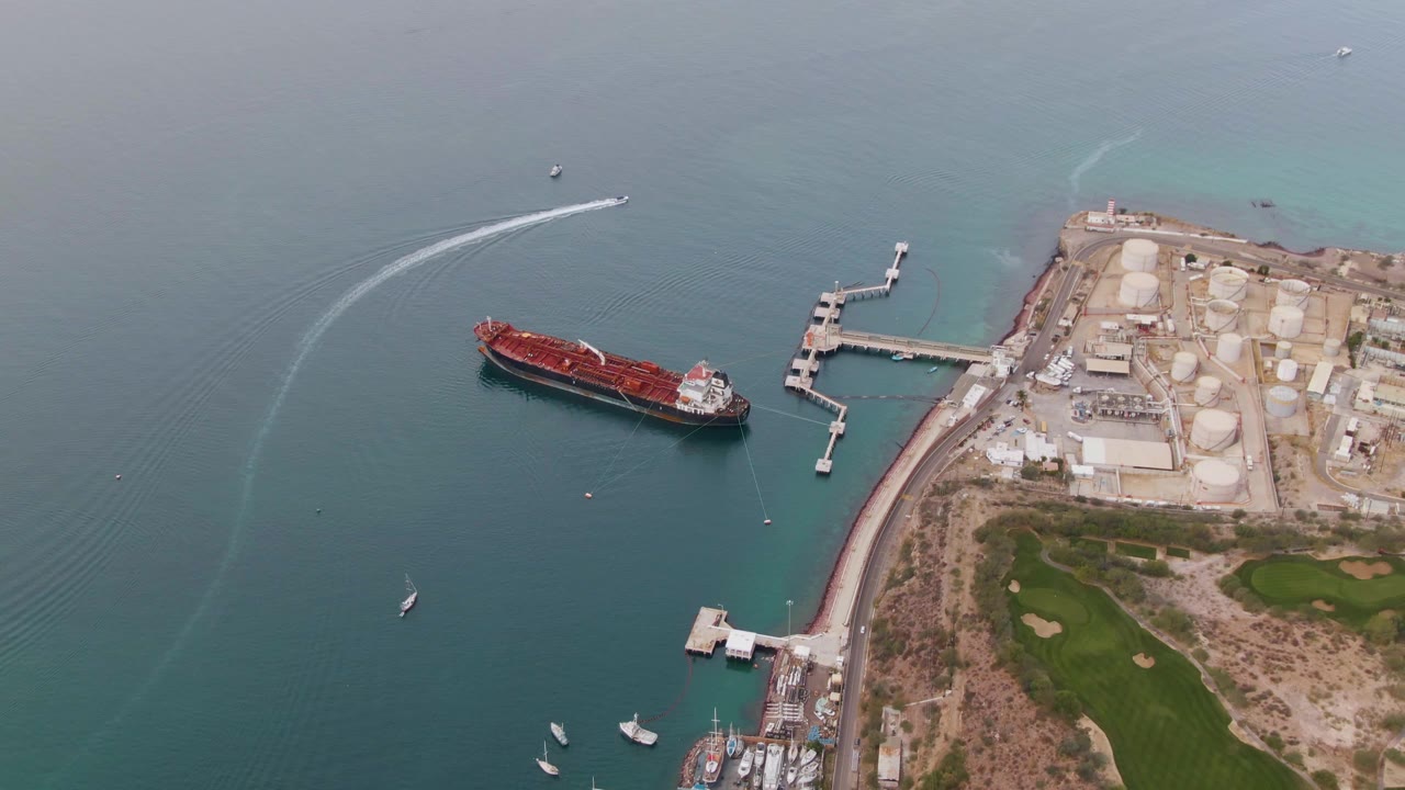 High aerial frame over La Paz, Baja California Sur showing the red cargo ship, angular industrial pier, marina boats below, golf course edge, blue-green water, and no visible people.