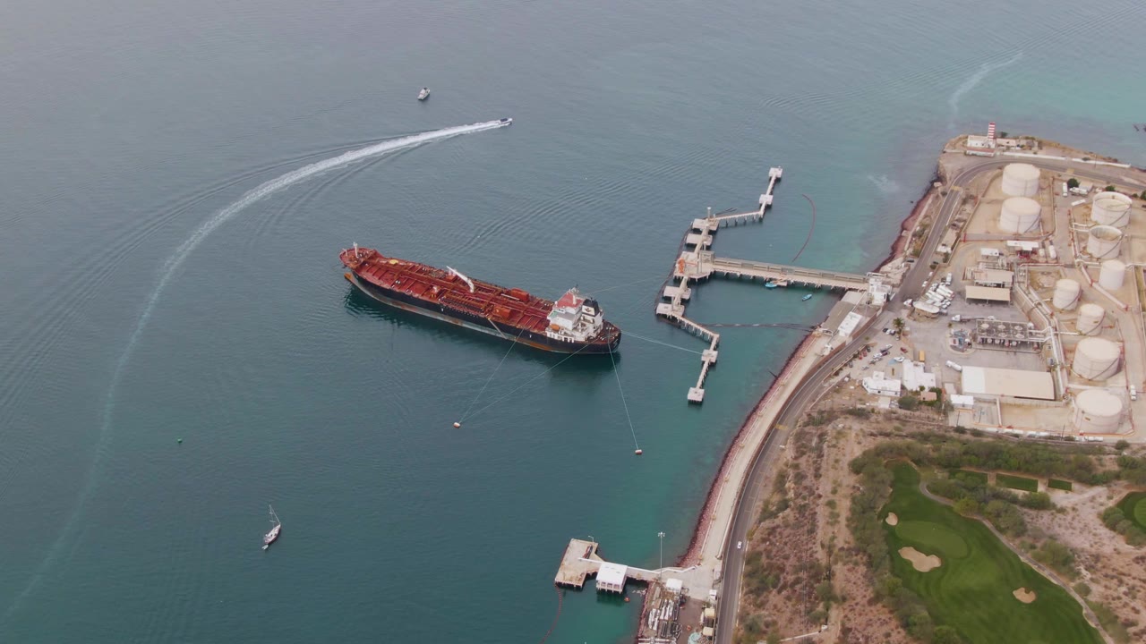 Closer aerial frame in La Paz, Baja California Sur with the red cargo ship beside the industrial pier, pale storage silos on shore, a small sailboat below, and no visible people.