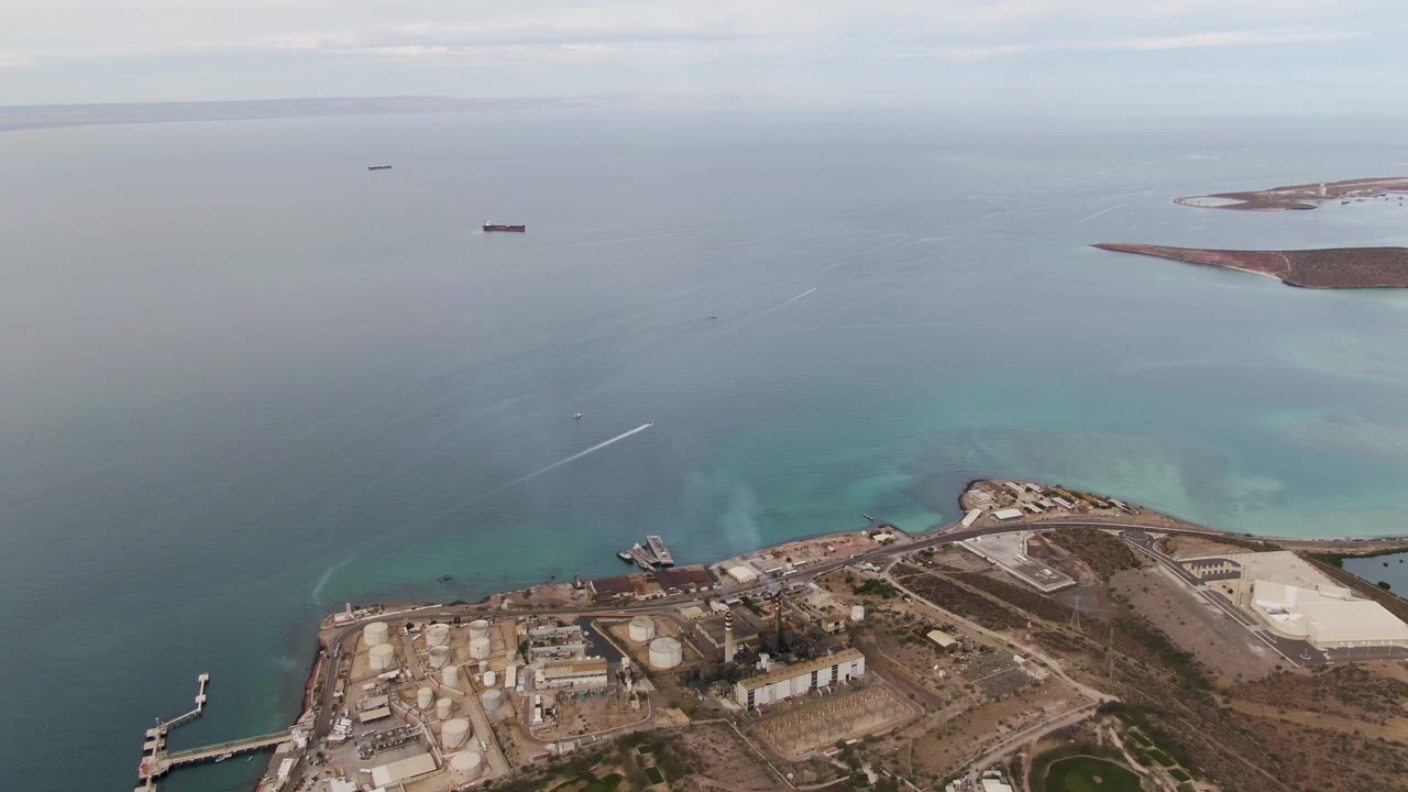 Aerial view of coastal industrial tanks, docks, turquoise water, and distant ships.