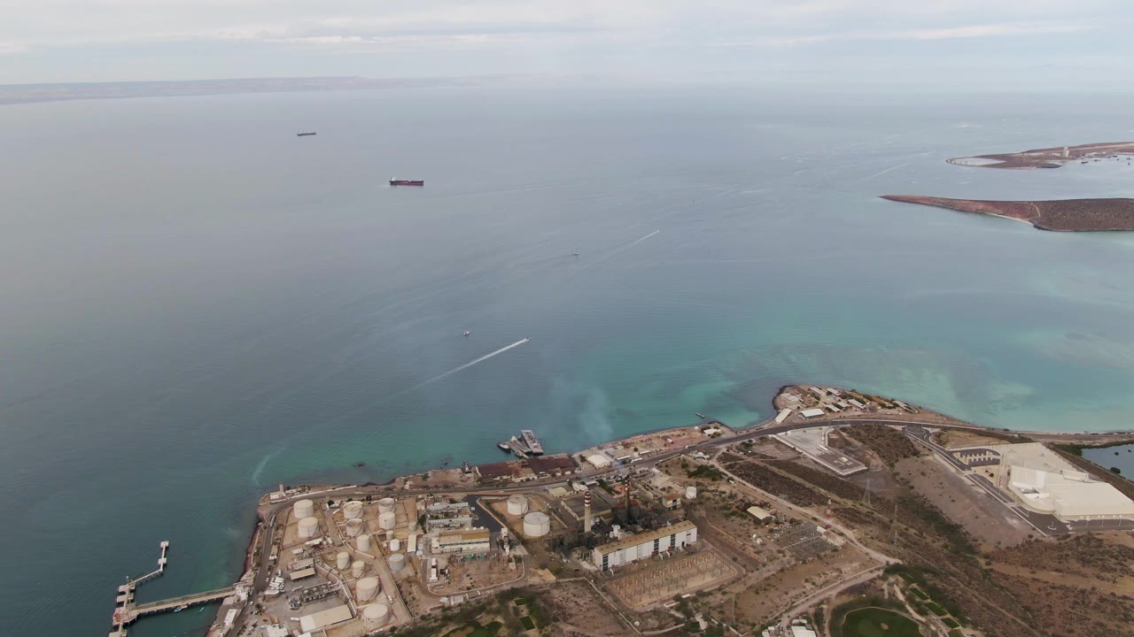 La Paz aerial view with waterfront tanks and dock structures in the lower frame, pale smoke rising near shore, and several ships spaced across the blue water, with no visible people.