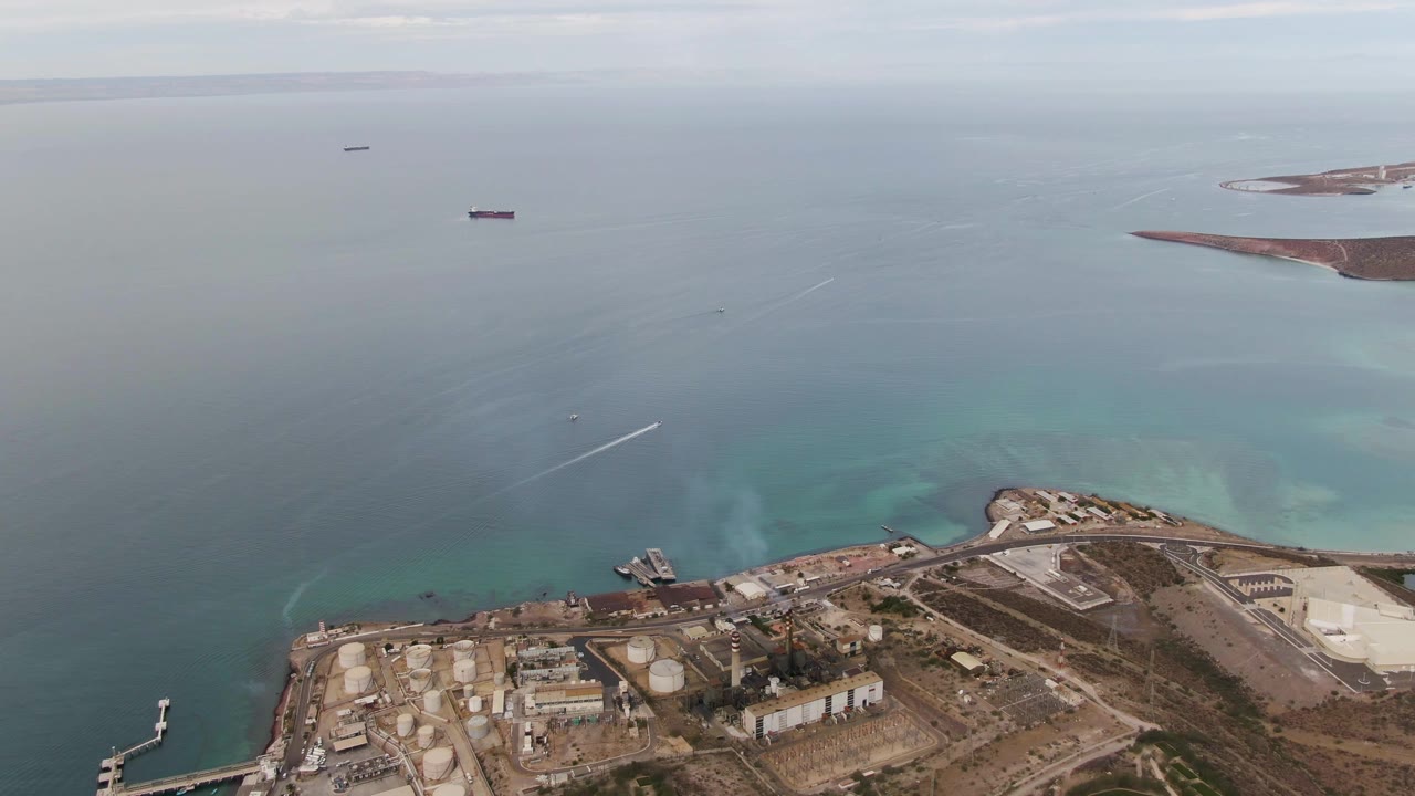 High drone frame of the La Paz coastal industrial site, with docks and tanks along the shore, turquoise water near land, deeper water beyond, distant ships, and no visible people.