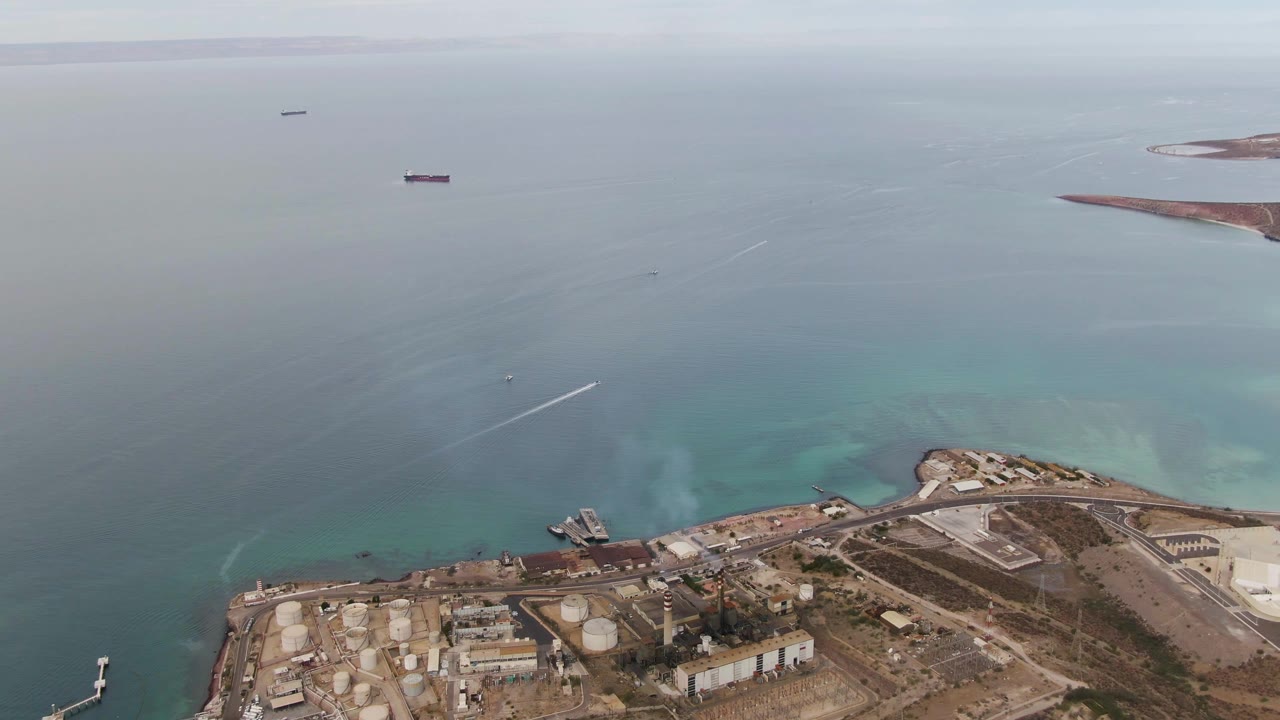 Aerial view in La Paz with the industrial shoreline lower in frame, a dock jutting into the water, anchored ships in the distance, a hazy horizon, and no visible people.