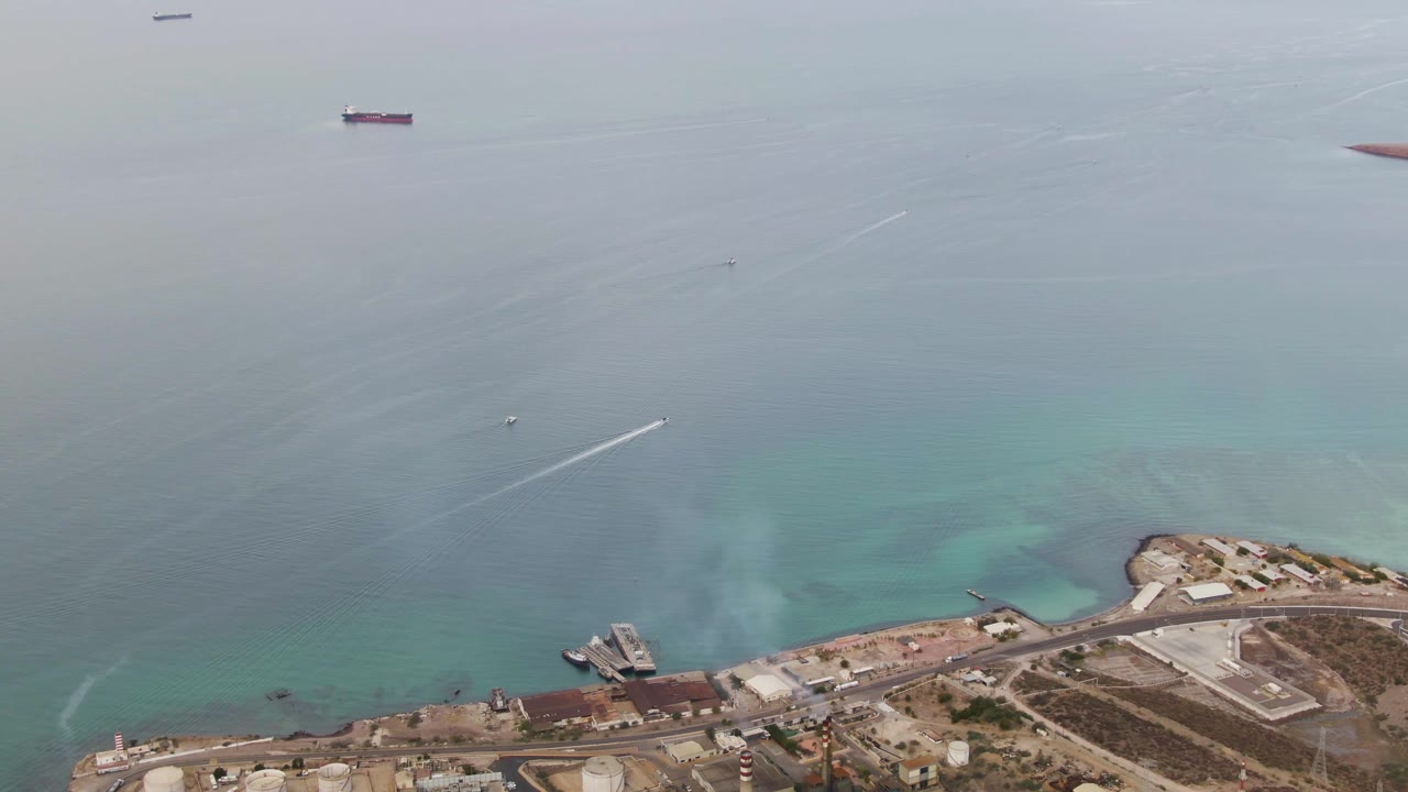 Aerial view in La Paz, Baja California Sur with the speedboat slightly farther across open water, its white wake stretching toward the industrial coast below and ships on the horizon, with no visible people.