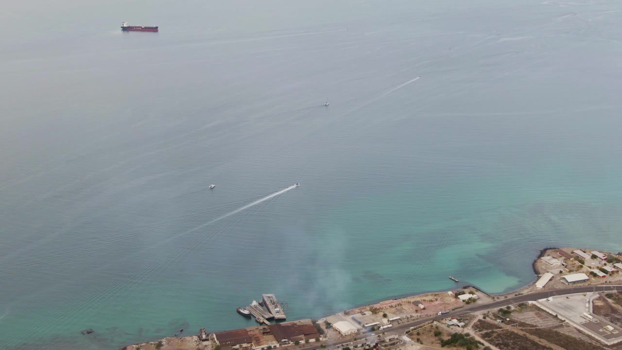 Aerial frame over La Paz where the speedboat and its wake sit over open blue water, while the lower edge shows port structures, a road, and shoreline tanks with no visible people.