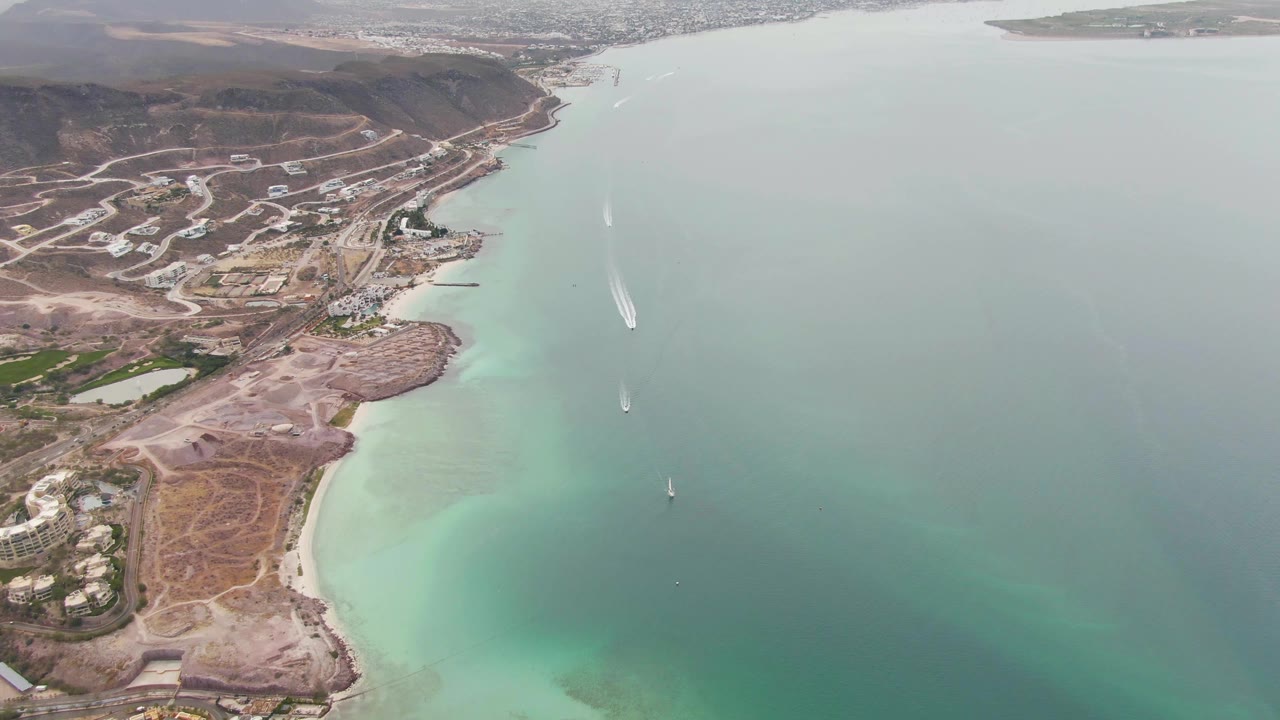 Drone view of coastal development beside blue-green water with two boat wakes offshore.