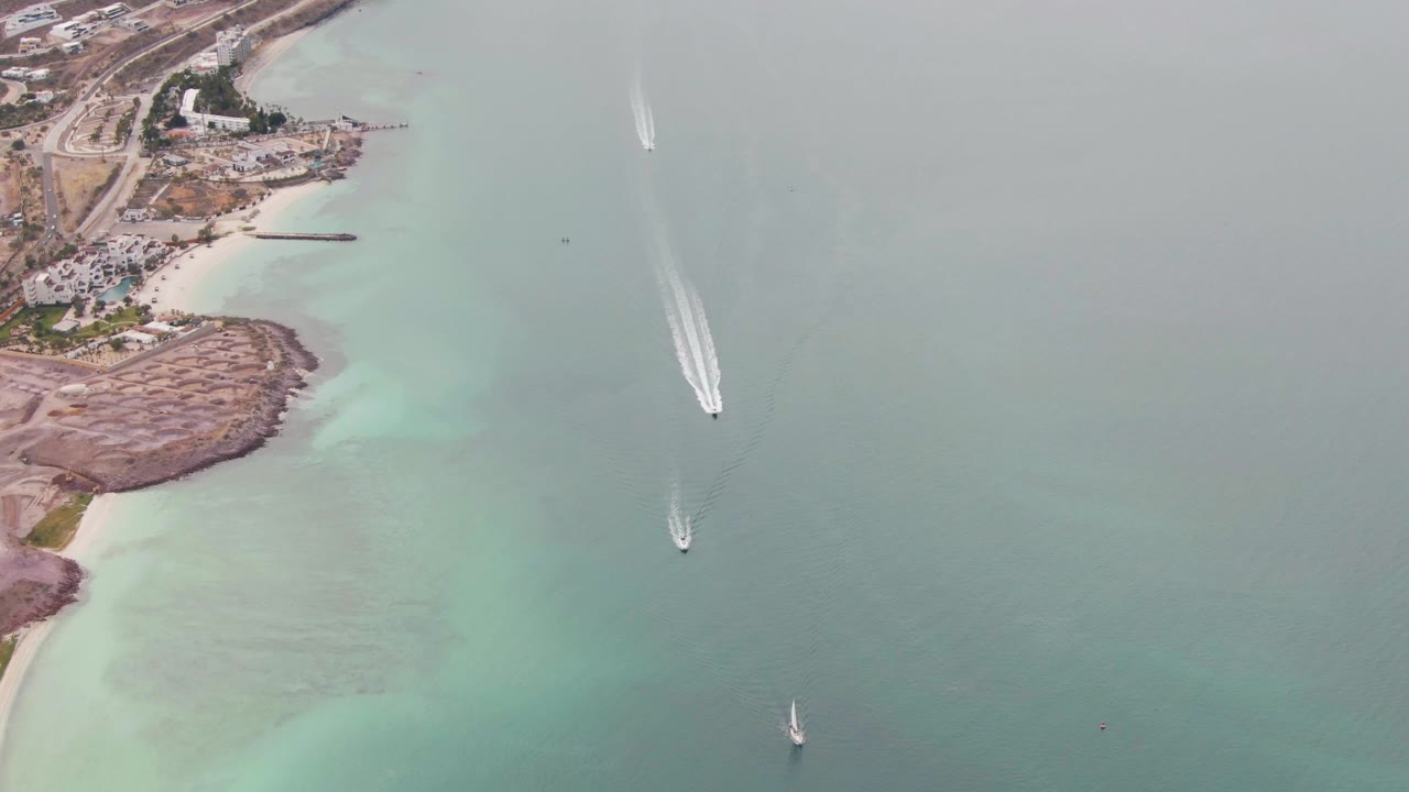 Aerial frame over La Paz, Baja California Sur shows turquoise shallow water beside a rocky and sandy shoreline, with coastal buildings, a road, two moving boats, a sailboat, and no visible people.