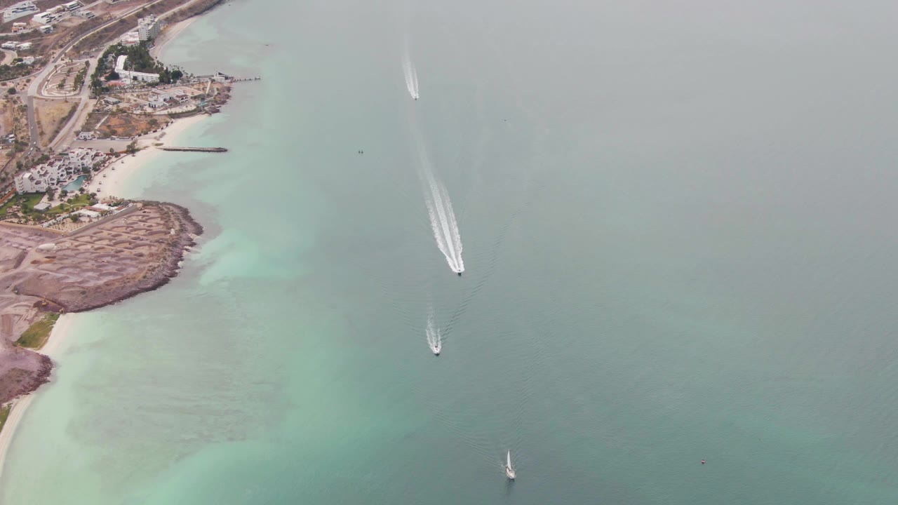 At 0:02, the drone-style view over La Paz shows the shoreline pushed farther left, open turquoise water filling most of the frame, a boat wake near the upper right, another boat near center, a sailboat lower right, and no visible people.