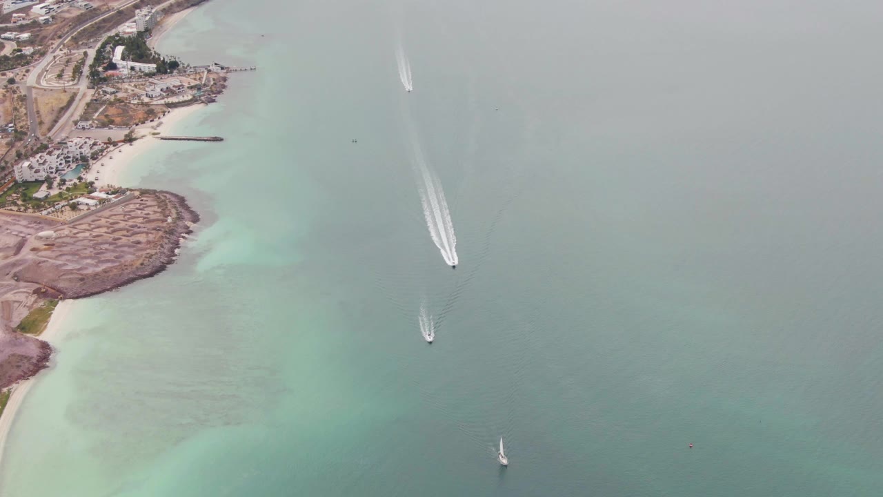Overhead coastal water scene with boats, wakes, rocky shore, beach, road, and buildings.
