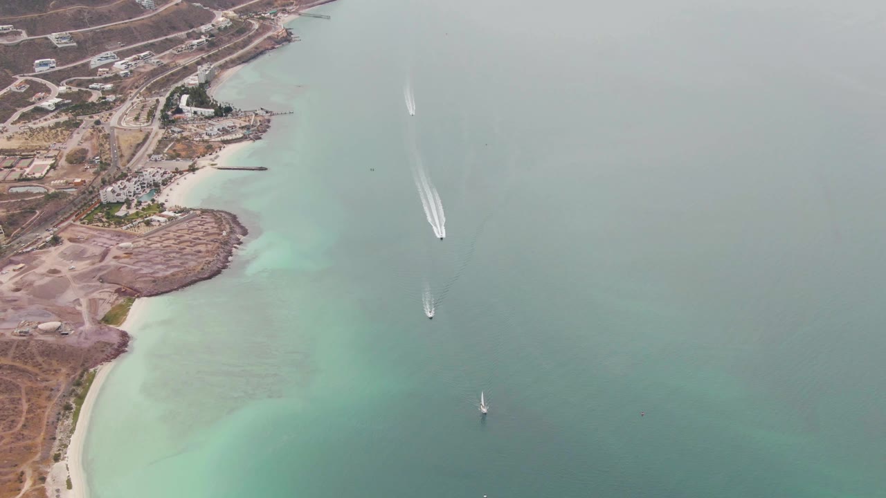 Aerial view in La Paz, Baja California Sur with the main white wake farther across open water, a sailboat lower in frame, shoreline development at left, and no visible people.