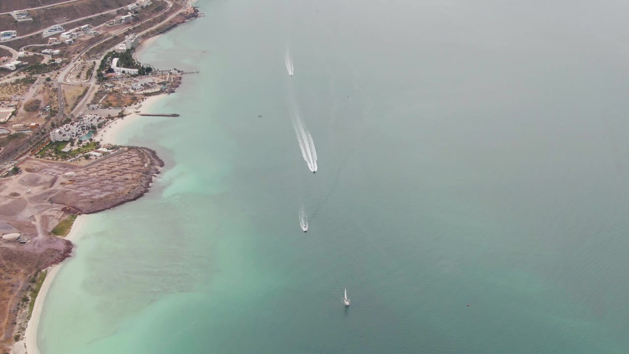 Aerial frame over the La Paz coast showing a bright wake near the upper water, a sailboat near center right, turquoise shallows along the beach, roads, buildings, and no visible people.