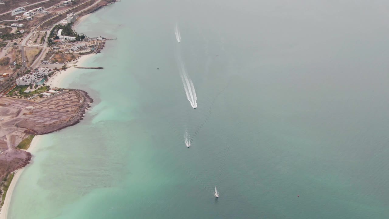 Aerial view of La Paz coastal water with a long white wake near the center, another small boat trail lower in frame, a sailboat near the bottom right, shoreline land at left, and no visible people.