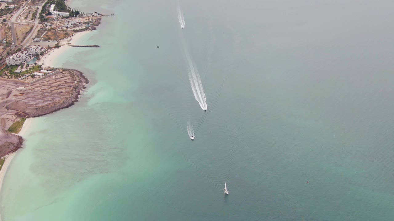 Aerial view of turquoise coastal water, rocky shoreline, boat wake, and small sailboats.