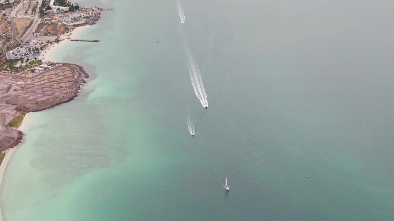 Aerial view over La Paz coastal water with the rocky point and pale beach on the left, a longer white wake in the upper center, a sailboat below it, another lower in frame, and no visible people.