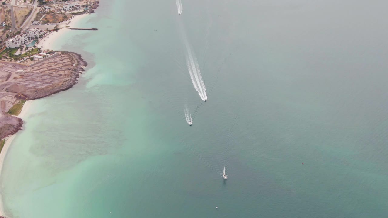 Aerial frame of the La Paz coast with the rocky shoreline shifted farther left, pale shallows along the beach, a white motorboat wake near the upper right area, two small sailboats below, and no visible people.
