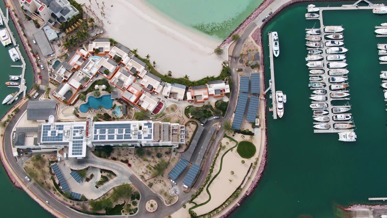 Top-down aerial frame over La Paz, Baja California Sur, showing a pale beach, waterfront residences, a blue pool, a curved road, marina slips, and no visible people.