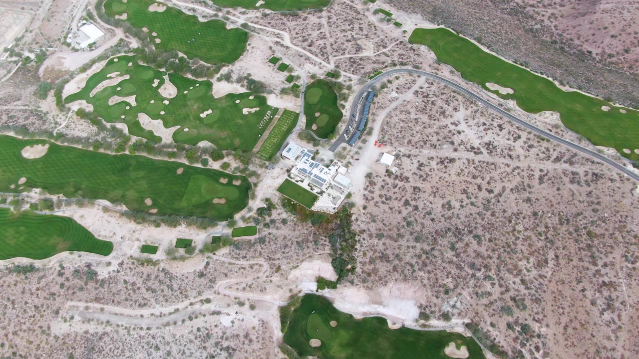Second aerial frame over the La Paz golf course, with the building cluster near the lower left, a curving road at center right, bright fairways, sand traps, and no visible people.