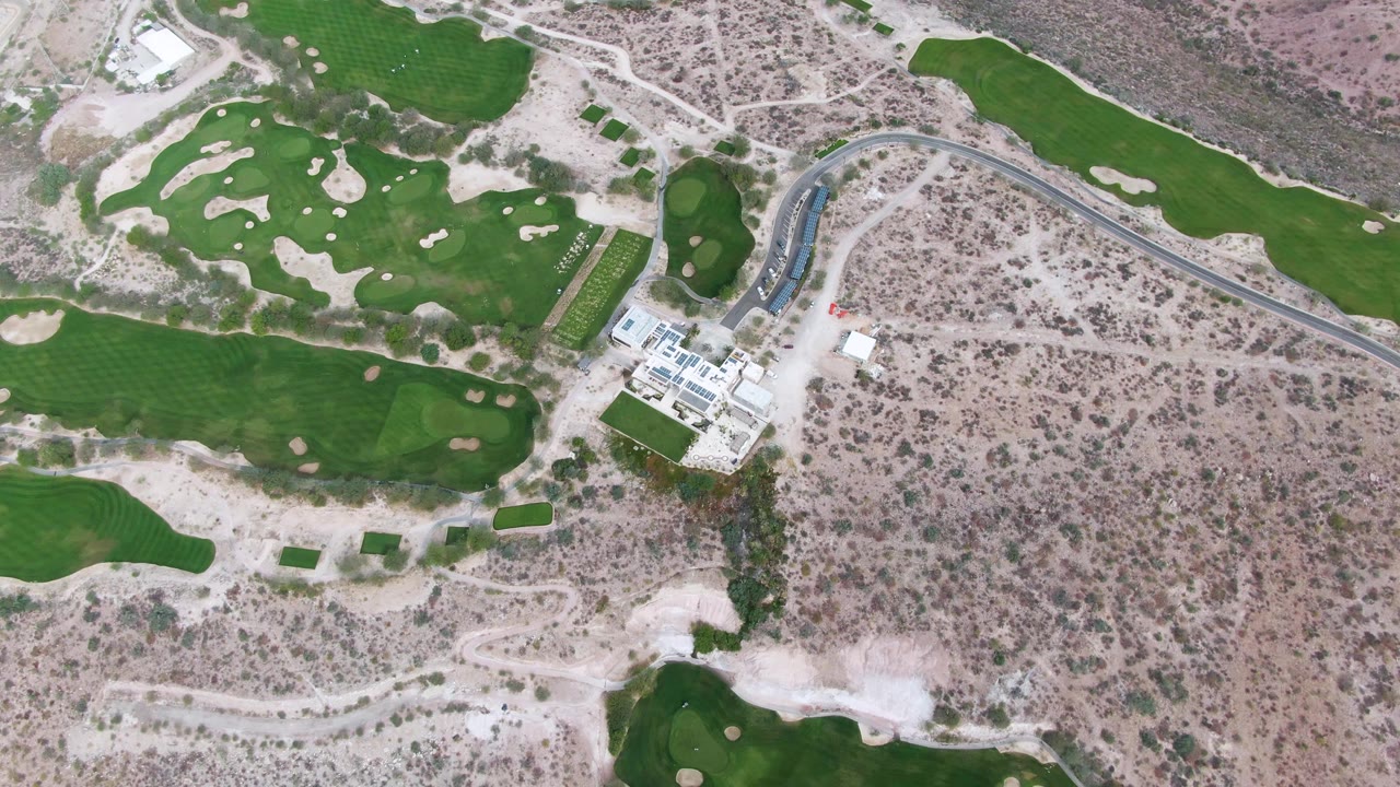 Aerial frame in La Paz, Baja California Sur, showing the course building left of center, a paved road curving through dry terrain, fairways above and below, sand traps, and no visible people.