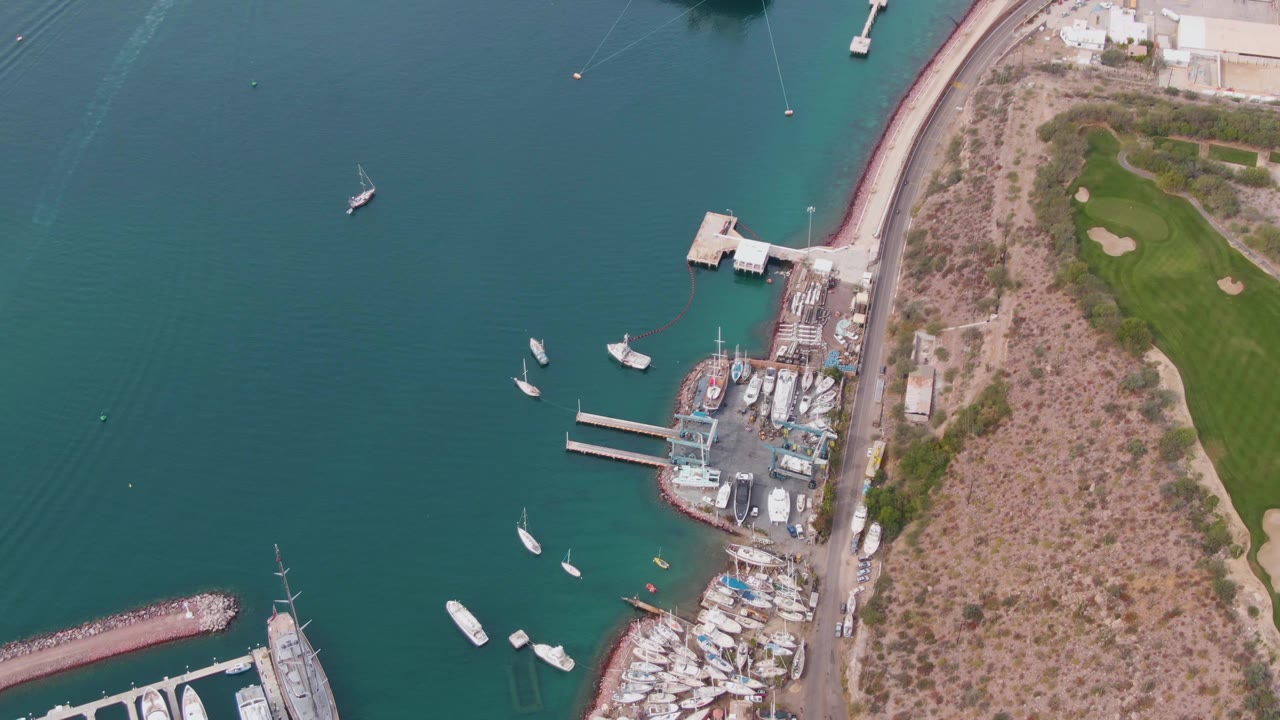 High aerial frame over La Paz harbor with teal water, scattered boats, marina docks, a shoreline road, scrubland, green golf course fairways, and no visible people.