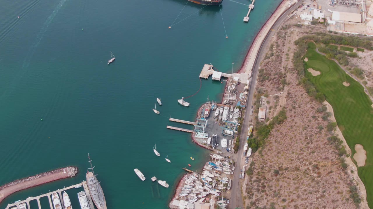 Aerial view of harbor boats beside docks, coastal road, scrubland, and golf course.