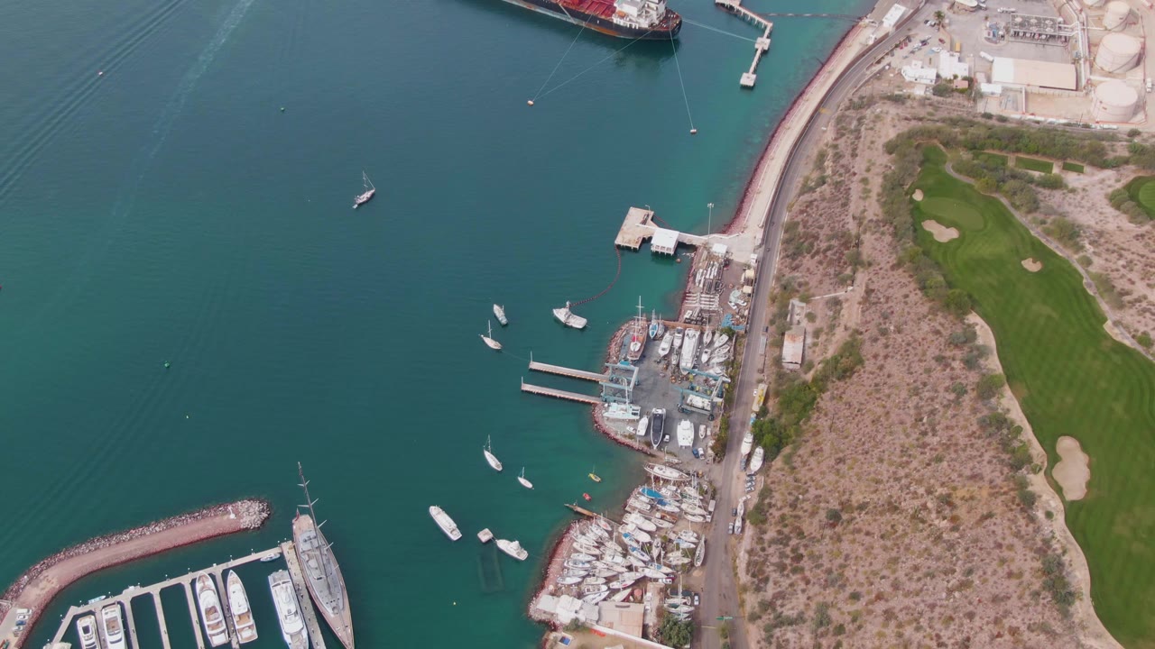 Aerial frame over La Paz harbor showing anchored boats, marina piers, a red cargo ship near the upper edge, a coastal road, dry scrubland, golf fairways, and no visible people.