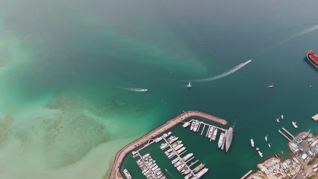 Overhead view in La Paz, Baja California Sur, showing turquoise harbor water, a marina breakwater at the lower edge, several boats, a curved speedboat wake near the top, and no visible people.