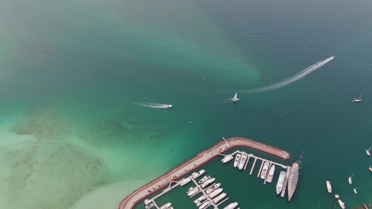 Aerial frame from La Paz, Baja California Sur, with the marina breakwater and docked boats at lower right, pale shallows at left, small boats on open water, visible wakes, and no visible people.