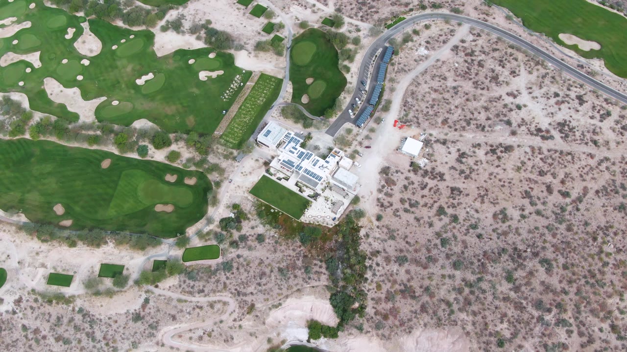 Overhead aerial frame in La Paz, Baja California Sur showing green golf course areas, pale sand traps, a white building complex, a curved road, dry brushland, and no visible people.